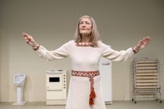 Ann Petrén i vit klänning med röda detaljer står med armarna utsträckta på en teaterscen.