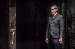 Lars Lind i kostym står på en mörk scen med en yxa synlig i bakgrunden.