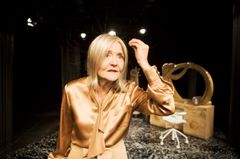 Ann Petrén i silkig blus på scen med rekvisita i bakgrunden.