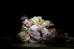 En person i en gyllene dräkt håller om en annan person som ligger ner. Scenen är mörkt belyst.