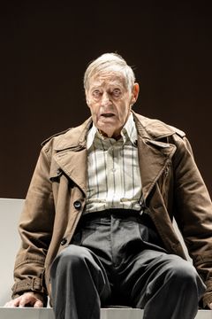 Lars Lind i en brun kappa och randig skjorta, sittande mot en mörk bakgrund.