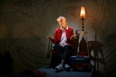 En äldre kvinna sitter i en fåtölj på ett podium, på väggen bakom henne anar man ritade porträtt.