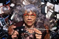 En äldre kvinna med långt, grått hår och röda glasögon ligger bland en massa kassettband och håller upp ett av banden framför sig.