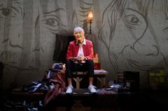 En äldre kvinna som sitter på ett podium bland massa kassettband. På väggen bakom henne ser man ritade porträtt.