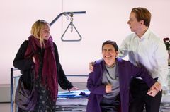 Tre skådespelare i en teaterscen står vid en sjukhussäng.