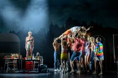 En scenbild från teaterföreställningen "Landet utom sig" med en gruppskulptur av skådespelare i ett dramatiskt ljus på scenen.