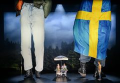 Scen från "Landet utom sig" med tre personer, en stor svensk flagga och förstärkta ben av figurer i förgrunden.