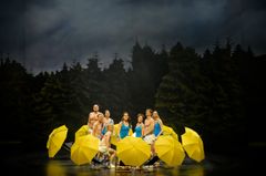 En grupp människor i scenkläder står omgivna av gula paraplyer, på en scen med en skogig bakgrund.