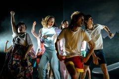 Grupp av skådespelare på scen som gestikulerar energiskt i en teaterproduktion.