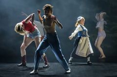 En grupp skådespelare dansar energiskt på en scen med en dimmig bakgrund.