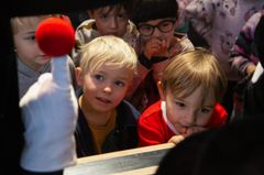 En grupp barn tittar fascinerat på en dockteaterföreställning med en vit handskklädd dockhand som håller i en röd boll.
