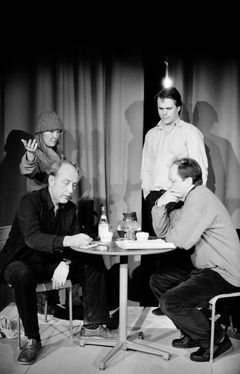 Två män sitter vid runt bord med dryck och anteckningar, två personer står bakom, varav en kvinna med mössa gestikulerar.