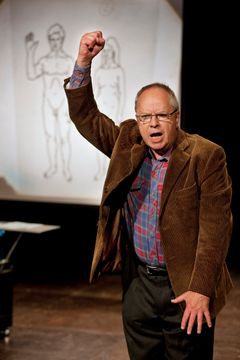 Helge Skoog i brun kavaj talar med höjd knuten näve framför teckning av nakna kroppar på blädderblock.