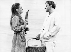 Gunnel Sporr i trenchcoat håller upp en gurka mot Helge Skoog i vit skjorta som bär en korg med bröd.