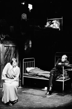 Kvinna sitter på stol och Helge Skoog sitter på sängkant i teaterscen med enkel sovrumsdekor.