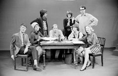 Åtta vuxna, fyra män och fyra kvinnor, sitter och står runt ett bord med papper och dryck.