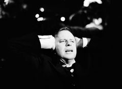 Helge Skoog i kostym sitter med händerna bakom huvudet, fotograferad i stark kontrast och närbild.