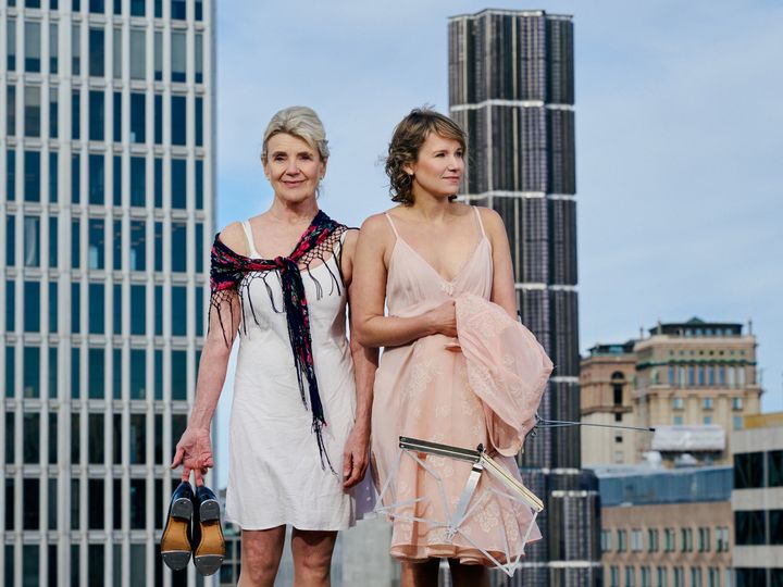 Stina Ekblad och Iiris Viljanen iklädda tunna klänningar och hållandes steppskor respektive ett notställ. De står utomhus på Kulturhuset Stadsteaterns tak.