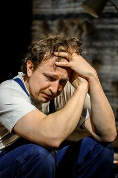 En person i blå overall med tankfull min håller huvudet i händerna. Mörk bakgrund.