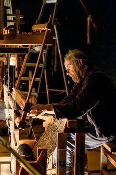 En man sitter vid en vävstol och väver i en mörk miljö.