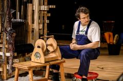 En man i arbetskläder sitter vid en vävstol på en teaterscen.