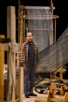 En person står framför en stor vävstol, omgiven av vävutsrustning.
