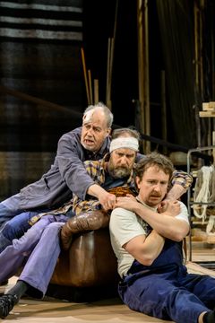 Tre män i arbetskläder sitter nära varandra på en scen med en teatral miljö.