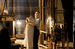 En man bär en rulle tyg på en teaterscen med vävstolar i bakgrunden.