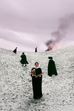 Fem personer klädda i mörka mantlar går mot en rökpelare på en snötäckt kulle. En person står i förgrunden med en bok.