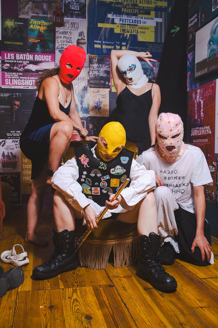 Fyra personer iförda balaklavor står mot en vägg fylld med affischer.