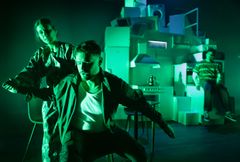 Två personer i förgrunden poserar dramatiskt i grönt ljus, omgivna av staplade hushållsapparater. En annan person sitter i bakgrunden.