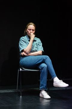 Kvinna sitter på en stol på scenen, klädd i jeans och en blå skjorta.