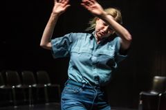 Kvinna i blå skjorta och jeans gestikulerar på en scen. Bakgrunden är mörk med några stolar.