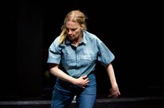 Kvinna i blå skjorta och byxor står på scen i en dramatisk pose.