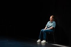 Kvinna i jeanskläder sitter på en stol på en mörk scen.