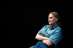 Kvinna i blå skjorta sitter med armarna korsade, mot en mörk bakgrund.