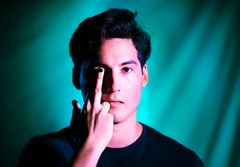 En person med mörkt hår visar ett finger framför ansiktet med en färgglad bakgrund.