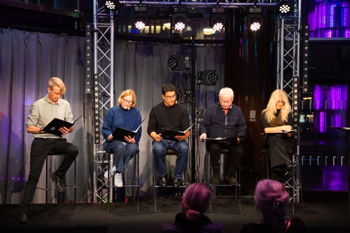En grupp människor sitter på stolar på en scen och läser från papper framför en liten publik.