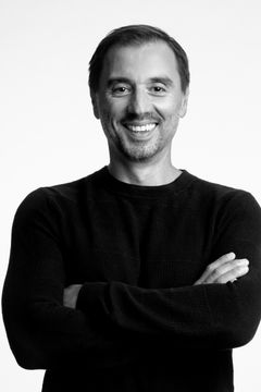 Bilden visar en leende person i svart tröja med armarna i kors, mot en vit bakgrund.