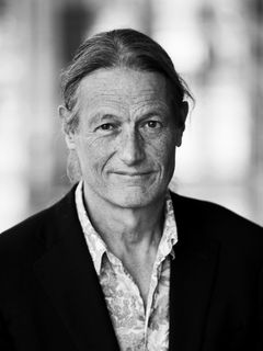 En man med långt hår och en kostym ser in i kameran. Svartvit bild.