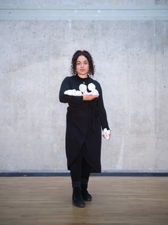 Yasmina Karli Malmsten med mörkt lockigt hår står mot en betongvägg och håller en modell av en skulptur i händerna.