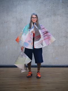 Anna Nyberg med långt grått hår, iklädd färgglad målarcanvas som en poncho och orange skor framför betongvägg.