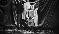 Fyra personer poserar i gammaldags kläder framför ett draperat tyg. En person sitter ner medan tre står bakom.