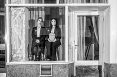 Helen Sjöholm och Anna Fahlstedt i ett tomt skyltfönster. De är svartklädda och sitter bredvid varandra på varsin pall, Anna håller en begravningsurna i handen.