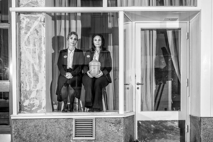 Helen Sjöholm och Anna Fahlstedt i ett tomt skyltfönster. De är svartklädda och sitter bredvid varandra på varsin pall, Anna håller en begravningsurna i handen.