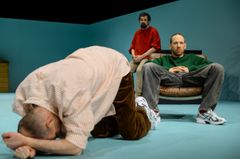 Tre personer på en scen i en teaterföreställning. En man ligger hopkrupen på golvet, en annan sitter i en stol, och den tredje står i bakgrunden.