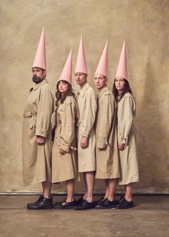 En grupp människor i trenchcoats och rosa strutformade hattar står på rad framför en neutral bakgrund.