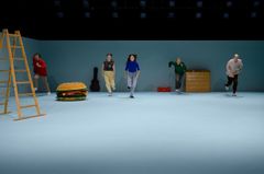 Ensemblen springer mot scenframkanten med rekvisita som stor hamburgare och stege.