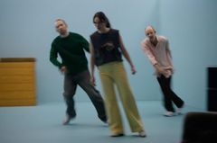 En scen med tre personer i rörelse på en teaterscen. De har olika kläder och bakgrunden är neutral.