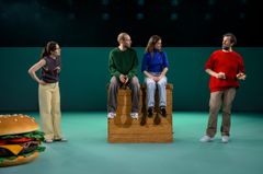 Fyra personer på scenen, tre sitter på en plint och en står. I förgrunden finns en stor hamburgermodel.